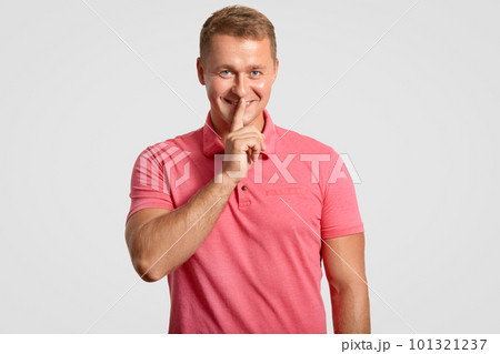 Waist up portrait of attractive man has appealing look, makes silence sign, dressed in casual pink t shirt, asks not spread private information, isolated over white background. Secrecy concept 101321237