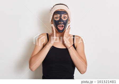 Winsome girl treating and caring for her skin. Woman with hairband on head and facial mask looking at camera over white background, keeping mouth opened, expressing astonishment. 101321481