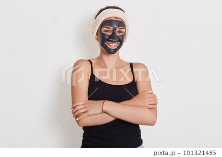 Close up portrait of young female applies homemade facial clay mask, has white hairband around head, smiles happily, keeps hands folded against white background. Close up portrait of young female applies homemade facial clay mask, has white hairband around head, smiles happily, keeps hands folded against white background. 101321485