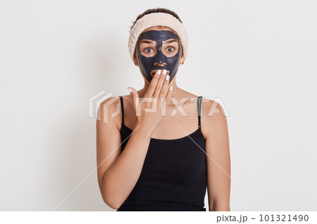 Emotional surprised Caucasian woman looks with shocked expression directly at camera, stands against white background, cleans her skin with mud cosmetic mask. 101321490