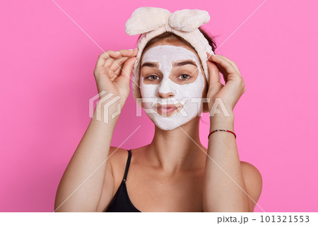 Young lady wearing sleeveless t shirt and headband, looking at camera with cosmetic mask on face, doing beauty procedures in morning at home. Young lady wearing sleeveless t shirt and headband, looking at camera with cosmetic mask on face, doing beauty procedures in morning at home. 101321553