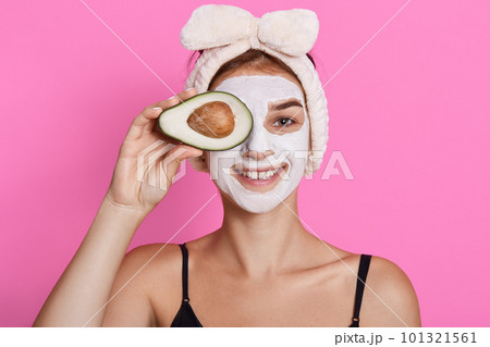 Attractive smiling female wearing sleeveless t shirt posing against rosy background, covering her eye with avocado, being in good mood, posing with moisturizing mask. 101321561