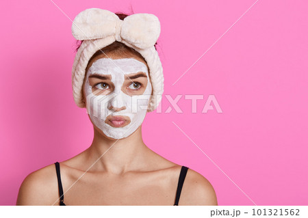 Woman with facial mask looking aside with pensive facial expression, having funny hairband, posing isolated over pink background. Copy space for advertisement. 101321562