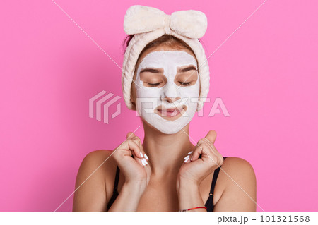 Woman with closed eyes and white facial mask on face, wearing sleeveless t shirt and hair band, standing against pink background, cosmetic procedures at home. 101321568
