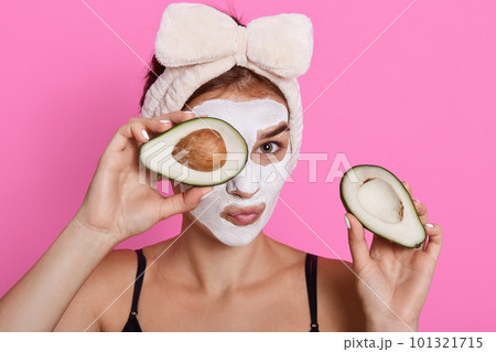 Beautiful spa woman with facial mask on face and holding halves of avocado in hands, looking at camera, doing cosmetology procedures at home, wearing hair band with bow. Beautiful spa woman with facial mask on face and holding halves of avocado in hands, looking at camera, doing cosmetology procedures at home, wearing hair band with bow. 101321715