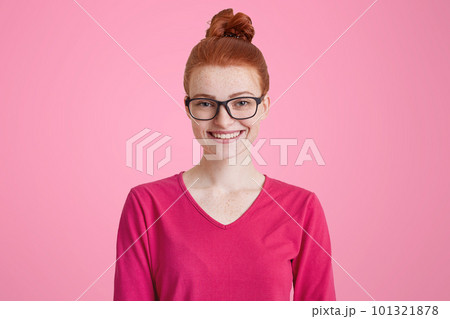 Isolated shot of happy ginger woman in pink sweater, wears square spectacles, has red hair tied in knot, freckled skin, pleasant smile, isolated over pink background. Cheerful female student indoor Isolated shot of happy ginger woman in pink sweater, wears square spectacles, has red hair tied in knot, freckled skin, pleasant smile, isolated over pink background. Cheerful female student indoor 101321878
