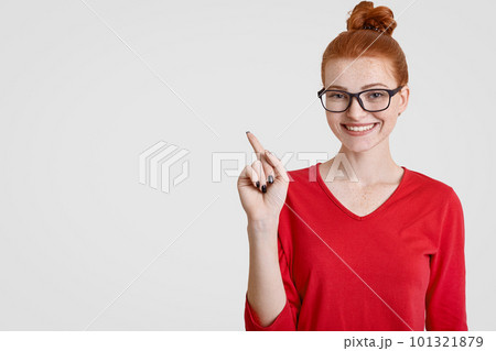 Intrigued ginger female teacher in spectacles attracts your attention to blank copy space, indicates with index finger, has charmig smile, likes students project work, poses over white background 101321879