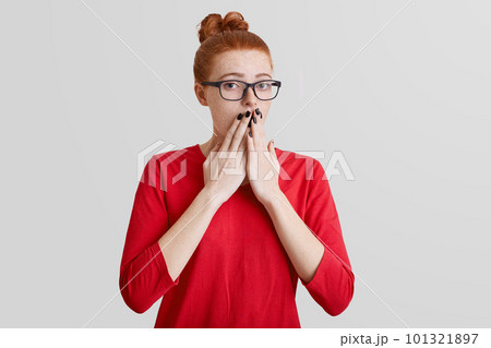 Startled frightened ginger woman covers mouth with hand, being afraid of something, wears spectacles and red sweater, poses alone against white background. Human reaction and feeling concept Startled frightened ginger woman covers mouth with hand, being afraid of something, wears spectacles and red sweater, poses alone against white background. Human reaction and feeling concept 101321897