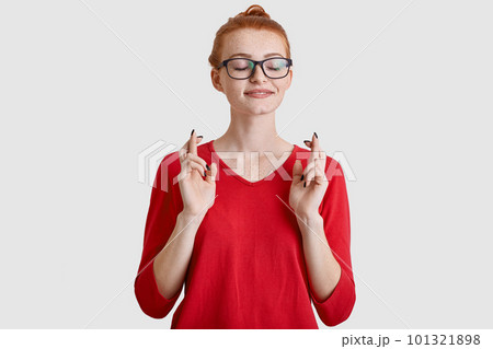 Freckled ginger young woman keeps fingers crossed, believes in good fortune, wears optical square spectacles, wears casual red jumper, hopes dreams come true, models against white background. Freckled ginger young woman keeps fingers crossed, believes in good fortune, wears optical square spectacles, wears casual red jumper, hopes dreams come true, models against white background. 101321898