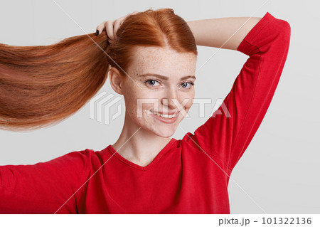 Portrait of happy redhead pretty young woman demonstrates her luxurious long hair, makes pony tail, going to have walk with friends, looks directly into camera, poses against white background. Portrait of happy redhead pretty young woman demonstrates her luxurious long hair, makes pony tail, going to have walk with friends, looks directly into camera, poses against white background. 101322136