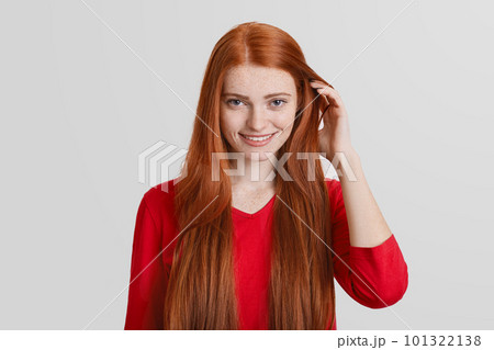 Portrait of redhead young female with long hair, has freckled face, pleasant smile, touches hair, isolated over white background. Ginger woman poses in studio, happy be photograhed. Portrait of redhead young female with long hair, has freckled face, pleasant smile, touches hair, isolated over white background. Ginger woman poses in studio, happy be photograhed. 101322138