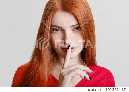 Close up portrait of freckled ginger young female holds index finger at lips, keeps silence, says Close up portrait of freckled ginger young female holds index finger at lips, keeps silence, says 101322147