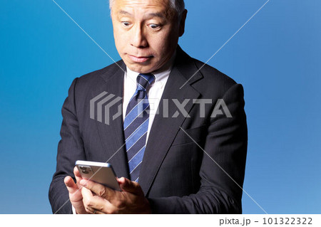 驚いた表情でスマホを確認するミドルシニアビジネスマン　ブルーバック　ビジネスイメージ 101322322