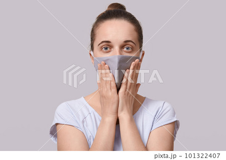 Close up picture of shocked frightened young lady putting hands close to face, wearing protective medical mask, opening eyes widely, being in panic, staying isolated at home. Coronavirus outbreak. 101322407