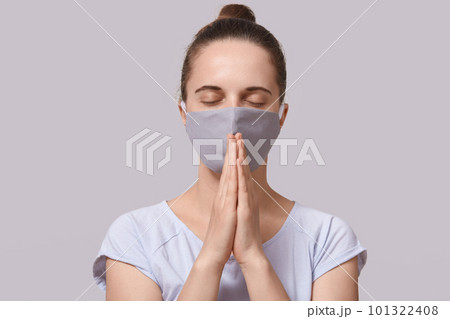 Picture of peaceful calm young woman being in quarantine, staying home, closing eyes, wearing antibacterial mask, sticking to rules of self isolation, putting hands together. Coronavirus concept. 101322408