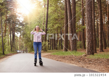 Attractive sporty woman wearing casual attire rollerblading and listening to music, keeping hands on her headphones, spending time with her family in forest. Winsome girl rinking in open air. Attractive sporty woman wearing casual attire rollerblading and listening to music, keeping hands on her headphones, spending time with her family in forest. Winsome girl rinking in open air. 101322454
