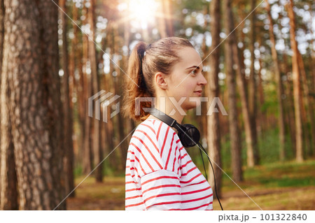 Profile of beautiful attractive young female looking ahead, having ponytail, chilling out in fresh air, being in forest, having walk, being delighted, having headphones around neck. Nature concept. 101322840
