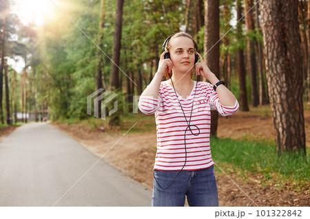 Horizontal photo of cheerful delighted good looking young female looking ahead, having headphones, listening to music, wearing jeans and sweatshirt, spending free time in peace. Rest concept. 101322842