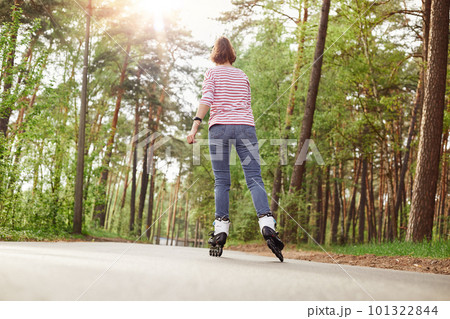Back view of skilled good looking young sportswoman having hobby, rollerskating, having rest, wearing striped sweatshirt and jeans, having training in forest, spending time in peace. Hobby concept. 101322844