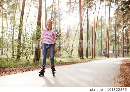Sporty energetic young female listening to music via headphones, having roller blades on legs, enjoying to spend time in park, having rest in open air. 101322876