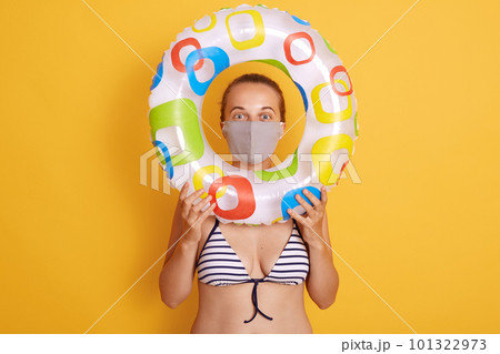Woman in striped swimsuit looking through rubber ring, wearing hygienic face mask to prevent virus on resort beach, rest and summer vacation with means of protection her health. Woman in striped swimsuit looking through rubber ring, wearing hygienic face mask to prevent virus on resort beach, rest and summer vacation with means of protection her health. 101322973