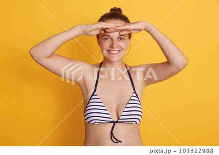 Beautiful woman wearing striped bikini swimwear posing isolated over yellow background, looks happy, stands smiling and looking far away with hand over head. Searching somebody. 101322988