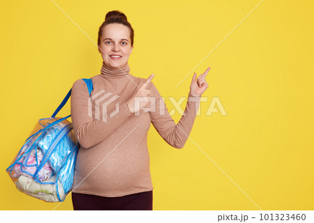 Young pretty pregnant woman pointing aside isolated over yellow background, looking smiling at camera, holds things for maternity hospital, being ready for giving birth. Copy space. 101323460