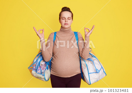 Nice adorable calm lovely beautiful expectant mom wearing casual attire, has hair bun, posing against yellow wall with bags for maternity hospital, trying to relax and not worry before giving birth. 101323481