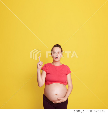 Surprised pregnant woman pointing finger up isolated over yellow background, lady with widely opened mouth touching her bare tummy, expectant mother. Copy space for add. 101323530