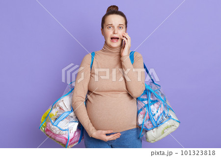 Portrait of worried pregnant lady, touching her belly and holding two bags with stuff, calling ambulance, keeps mouth opened, panic, asking for help, posing isolated over lilac background. 101323818