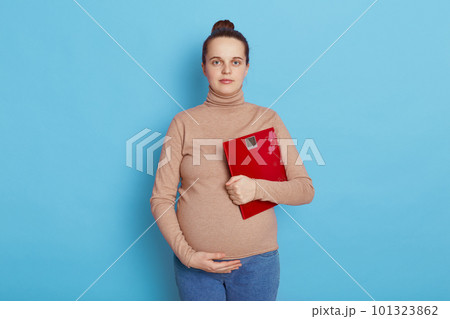 Pretty young pregnant woman holding floor scales in hands, future mother with hair bun touching belly and looking directly at camera, expectant mommy against blue wall. 101323862