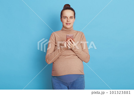 European pregnant with with pleasant appearance, holding phone in hands and looking directly at camera, needs to make call, wearing casually, isolated over blue background. European pregnant with with pleasant appearance, holding phone in hands and looking directly at camera, needs to make call, wearing casually, isolated over blue background. 101323881