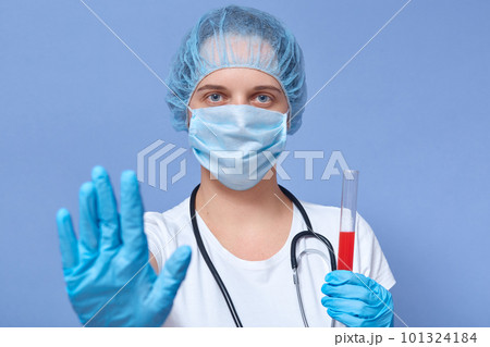 Picture of serious hard working doctor holding tube with blood for coronavirus test, making gesture, raising hand, wearing protective mask, hat and gloves, controling situation. Covid19 concept. Picture of serious hard working doctor holding tube with blood for coronavirus test, making gesture, raising hand, wearing protective mask, hat and gloves, controling situation. Covid19 concept. 101324184