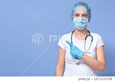 Indoor shot of doctor woman isolated over blue background, wears white shirt, medical sterile face mask, disposable hat, points aside with fore finger. Healthcare, medicine, health concept. Copy space 101324199