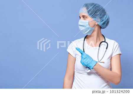 Picture of Caucasian doctor woman isolated over blue studio background, wearing hat, gloves and flumask, pointing aside with her forefinger, looking aside. Copy space for advertisment or promotion. Picture of Caucasian doctor woman isolated over blue studio background, wearing hat, gloves and flumask, pointing aside with her forefinger, looking aside. Copy space for advertisment or promotion. 101324200