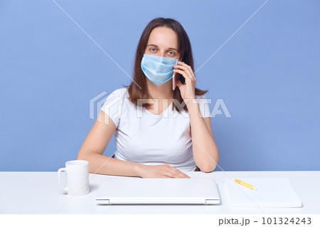 Portrait of good looking confident businesswoman looking directly at camera, talking over phone, holding smartphone, wearing antibacterial mask, working as freelancer. Work and quarantine concept. 101324243