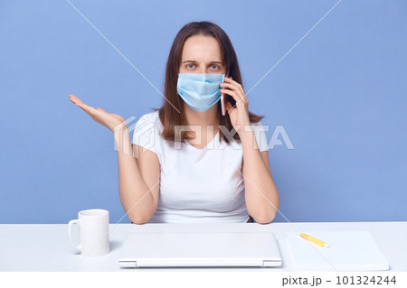 Image of woman wearing medical mask working from home, online freelance during quarantine, female talking via mobile phone and spreading hand aside, marketer discuding important questions remotely. 101324244