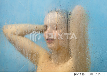 Healthy young female poses under matte blurred glass with water drops, takes shower in shower cabine, feels rejuvenation and enjoys hot water, has healthy soft skin after applying gel 101324599