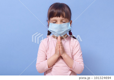 Coronavirus, self isolation, quarantine concept. Little Caucasian girl wearing mask for protect fron covid 19, standing with closed eyes and praying for stop corona virus, poses against blue wall. 101324694