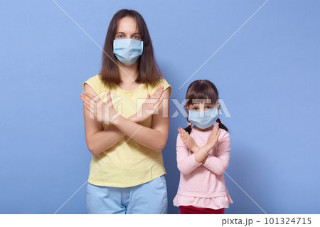 Image of mother and small girl with pigtails wearing medical protective masks against viral diseases during coronavirus epidemic, showing stop gesture with crossesd arms against blue background. 101324715