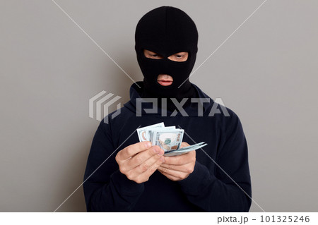 Swindler thief man counts stolen money, wears a black sweater, poses with a disguised face, a criminal breaks the law and takes money from other people, isolated over gray background. 101325246