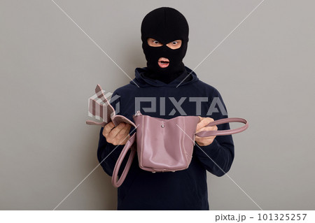 Young male criminal with stolen bag in his hands, looks in inside with shocked expression on his face, being very surprised that he sees in bag, isolated over gray background. Young male criminal with stolen bag in his hands, looks in inside with shocked expression on his face, being very surprised that he sees in bag, isolated over gray background. 101325257