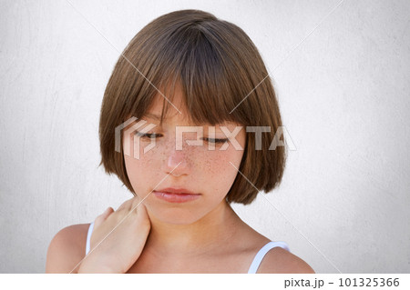 Close up of attractive little child with freckles and dark short hair keeping her hand on neck, looking seriously down, having thoughtful expression while posing against white concerete wall 101325366