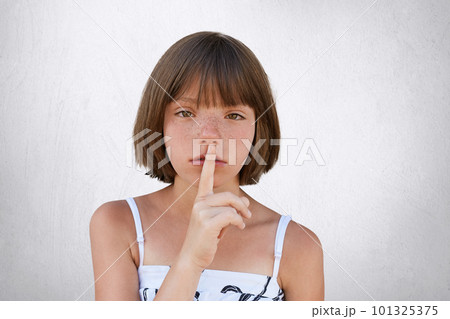 Be silent, hush! Adorable small kid showing silent sign asking to be noiseless as her little sister is sleeping. Freckled girl with stylish hairdo keeping her hand on mouth, showing hush sign. 101325375