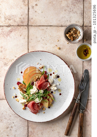 Salad of arugula and prosciutto or ham with the addition of sliced apple, cheese, walnuts and spices on a plate with cutlery on pink tile background. Top view, text space. Salad of arugula and prosciutto or ham with the addition of sliced apple, cheese, walnuts and spices on a plate with cutlery on pink tile background. Top view, text space. 101325696