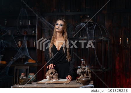 Halloween concept. Witch portrait close up with dreadlocks dressed black hood standing dark room with cage on background 101329228