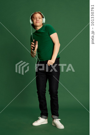 Exciting young man wearing headphones listening music and dancing over green background. Favourite melody Exciting young man wearing headphones listening music and dancing over green background. Favourite melody 101330761