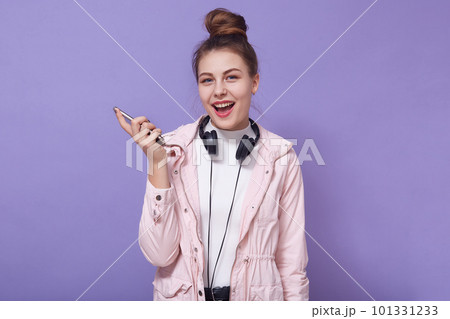 Happy excited woman wearing casual clothing,having headphones around neck, holding modern smart phone in hands, posing with opened mouth against lilac wall, adorable lady enjoying listening to music. Happy excited woman wearing casual clothing,having headphones around neck, holding modern smart phone in hands, posing with opened mouth against lilac wall, adorable lady enjoying listening to music. 101331233