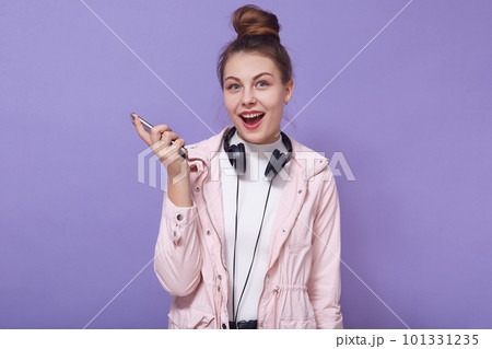 Portrait of charming lovely sweet good looking female opening mouth widely, holding smartphone, having headphones, listening to music, looking directly at camera. People and emotions concept. Portrait of charming lovely sweet good looking female opening mouth widely, holding smartphone, having headphones, listening to music, looking directly at camera. People and emotions concept. 101331235