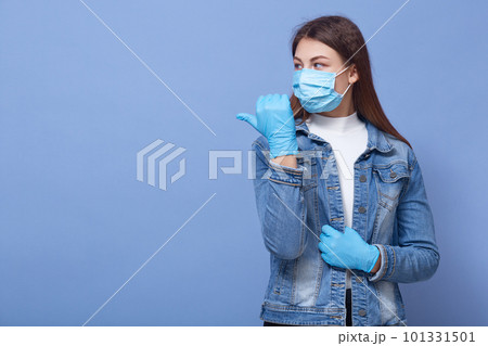Close up portrait of beautiful and young woman wearing white shirt and denim jacket, blue rubber gloves and flumask on face, pointing aside with thumb, isolated over blue background, copy space. 101331501